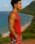 Man in a red tank top and floral tropical shorts standing on a hill overlooking a beach with mountains.
