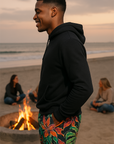 Man in black hoodie and colorful shorts standing by a beach fire pit with people sitting around.