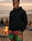 Man wearing a black hoodie and colorful shorts standing on a beach with a fire pit and people in the background.