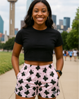 Woman wearing a black crop top and pink shorts with black floral patterns, standing in front of a city skyline.