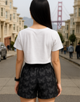 Woman in a white crop top and black butterfly patterned shorts standing on a city street.