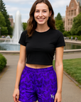 Woman wearing a black crop top and purple shorts with black butterflies, standing outdoors with a fountain and building in the background.
