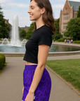 Woman wearing a black crop top and purple shorts with black butterflies, standing outdoors with a fountain and building in the background.