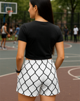 Woman standing on a basketball court wearing a black crop top and white shorts with black chain link pattern.