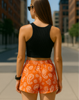 Woman standing on city street wearing RF-Wear Women's Paisley Shorts - Orange /White - Back View