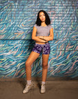 Woman standing near artful wall wearing RF-Wear Women's Paisley Shorts - Purple/White - Front View
