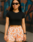 Woman wearing a black crop top and RF-Wear white/orange paisley shorts standing in front of a graffiti-covered wall.