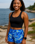 Woman in blue patterned shorts and black top standing on a path by the ocean with people in the background.