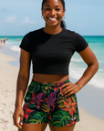 Woman in black cropped top and colorful shorts standing on the beach.