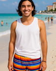 Man in blue top and colorful shorts standing on a beach with ocean and people in the background