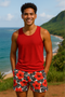 Man wearing a red tank top and floral tropical  shorts standing in a scenic location with ocean and mountains.