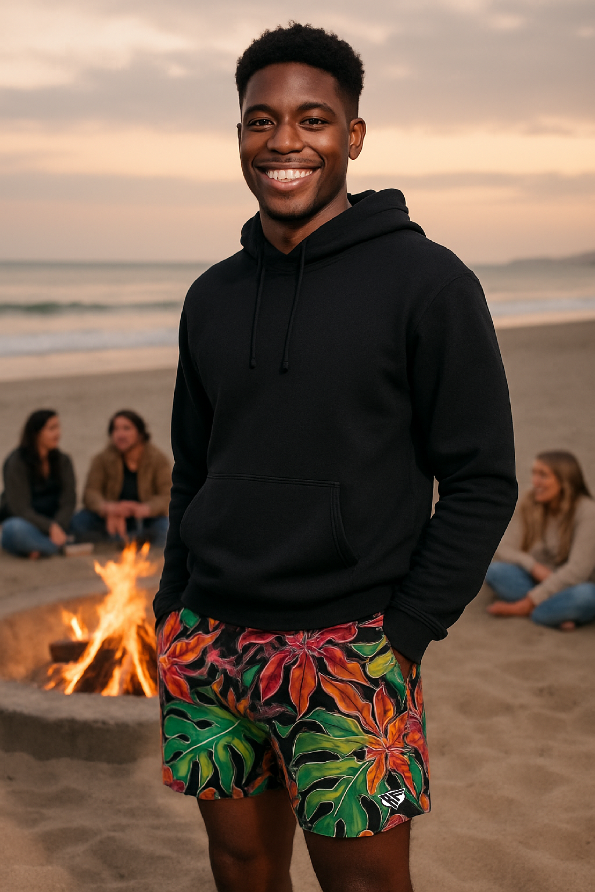 Man wearing a black hoodie and colorful shorts standing on a beach with a fire pit and people in the background.