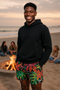 Man wearing a black hoodie and colorful shorts standing on a beach with a fire pit and people in the background.
