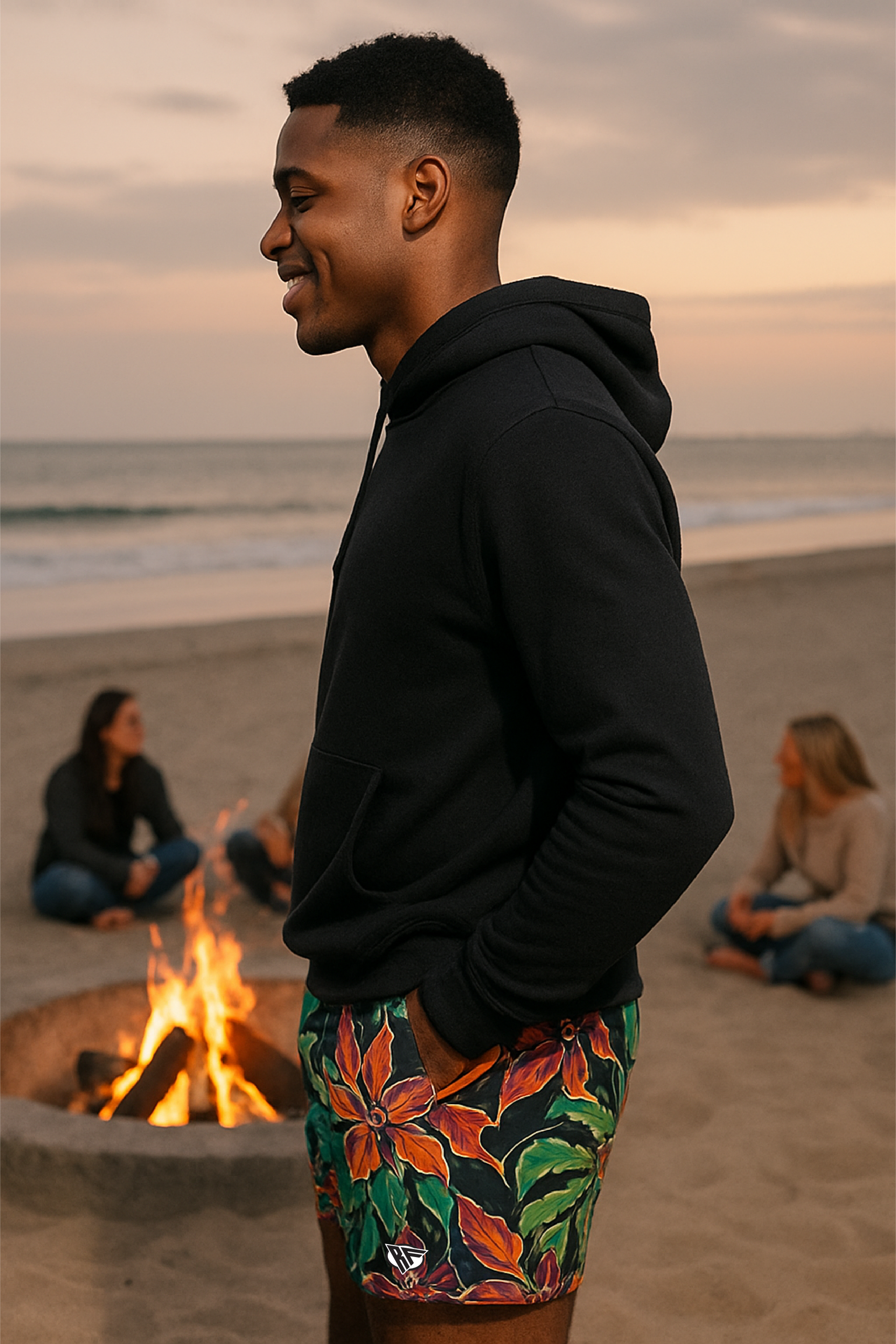 Man in black hoodie and colorful shorts standing by a beach fire pit with people sitting around.