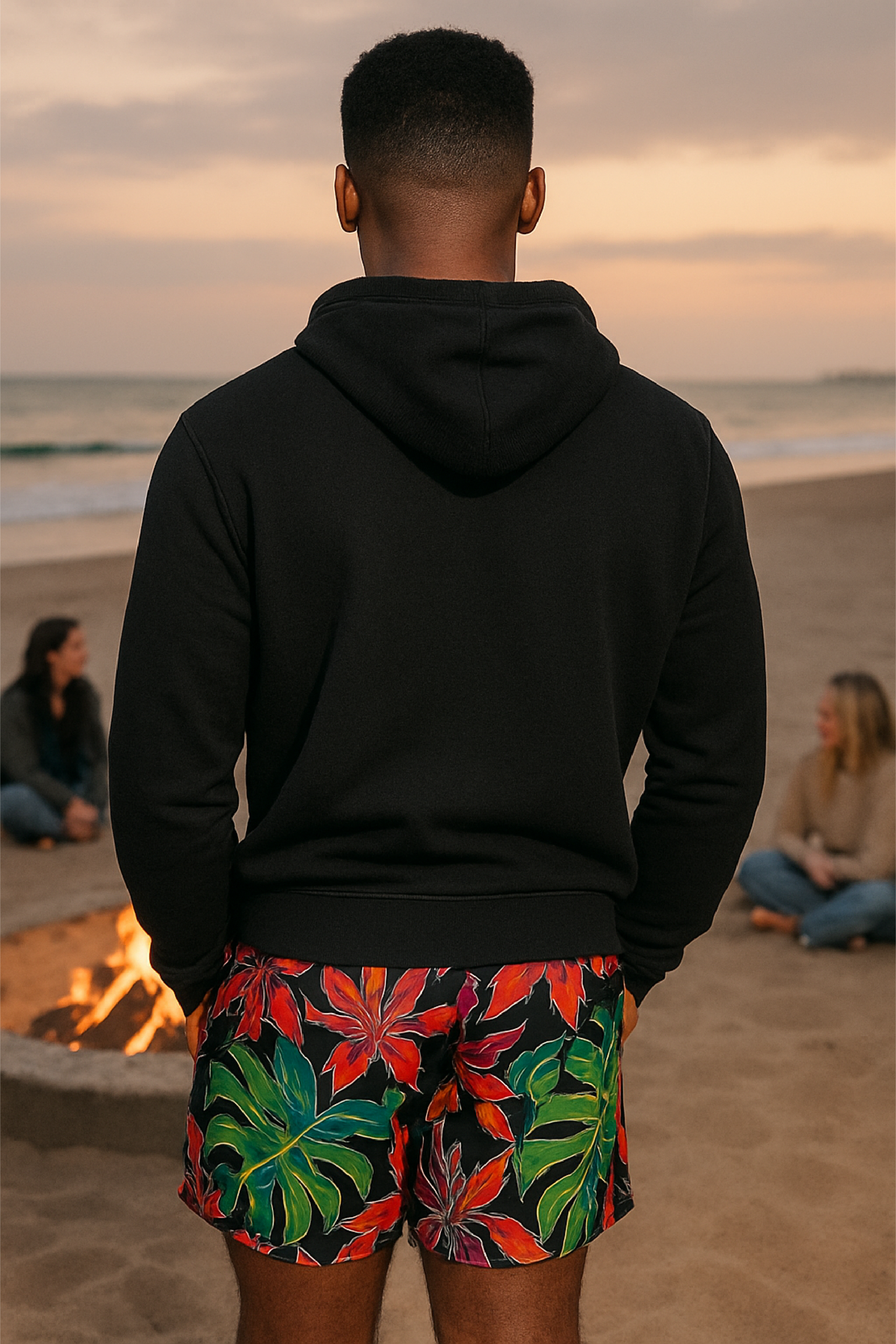 Man wearing a black hoodie and colorful shorts on a beach with people and a fire pit in the background.