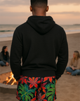 Man wearing a black hoodie and colorful shorts on a beach with people and a fire pit in the background.