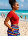Woman in a red top and colorful striped shorts standing on a beach with ocean and people in the background.