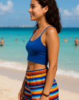 Woman in blue top and colorful striped shorts standing on a beach with ocean and sky in the background.