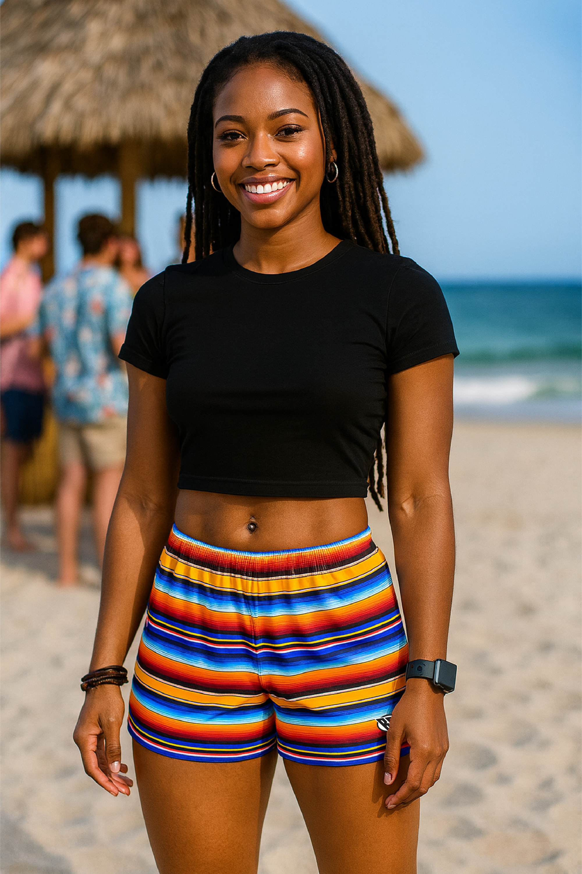 Woman in black top and colorful striped shorts standing on a beach with people and ocean in the background.