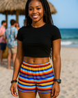 Woman in black top and colorful striped shorts standing on a beach with people and ocean in the background.