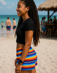 Woman in a colorful skirt and black top on a beach with people in the background