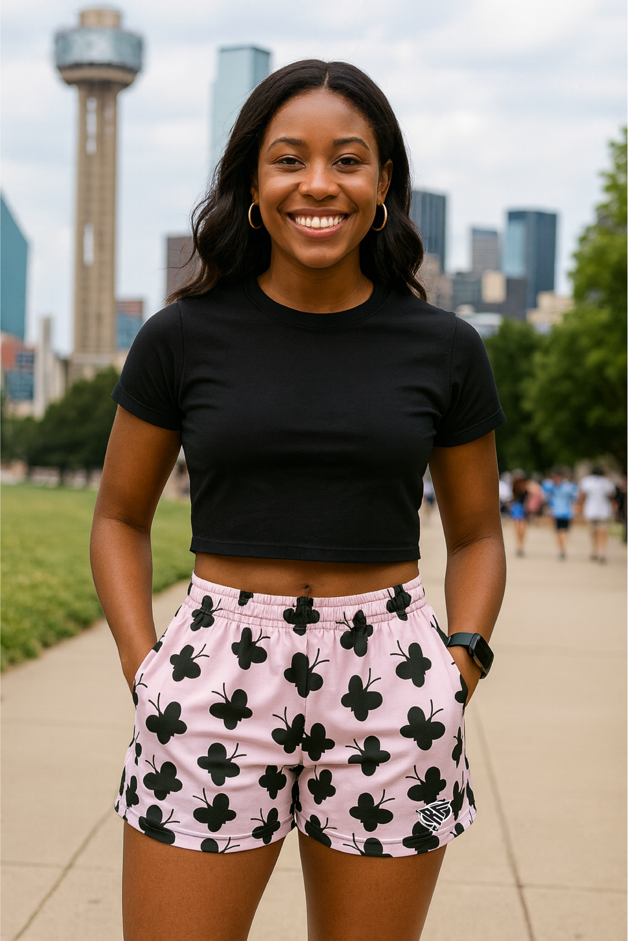 Woman wearing a black crop top and pink shorts with black floral patterns, standing in front of a city skyline.