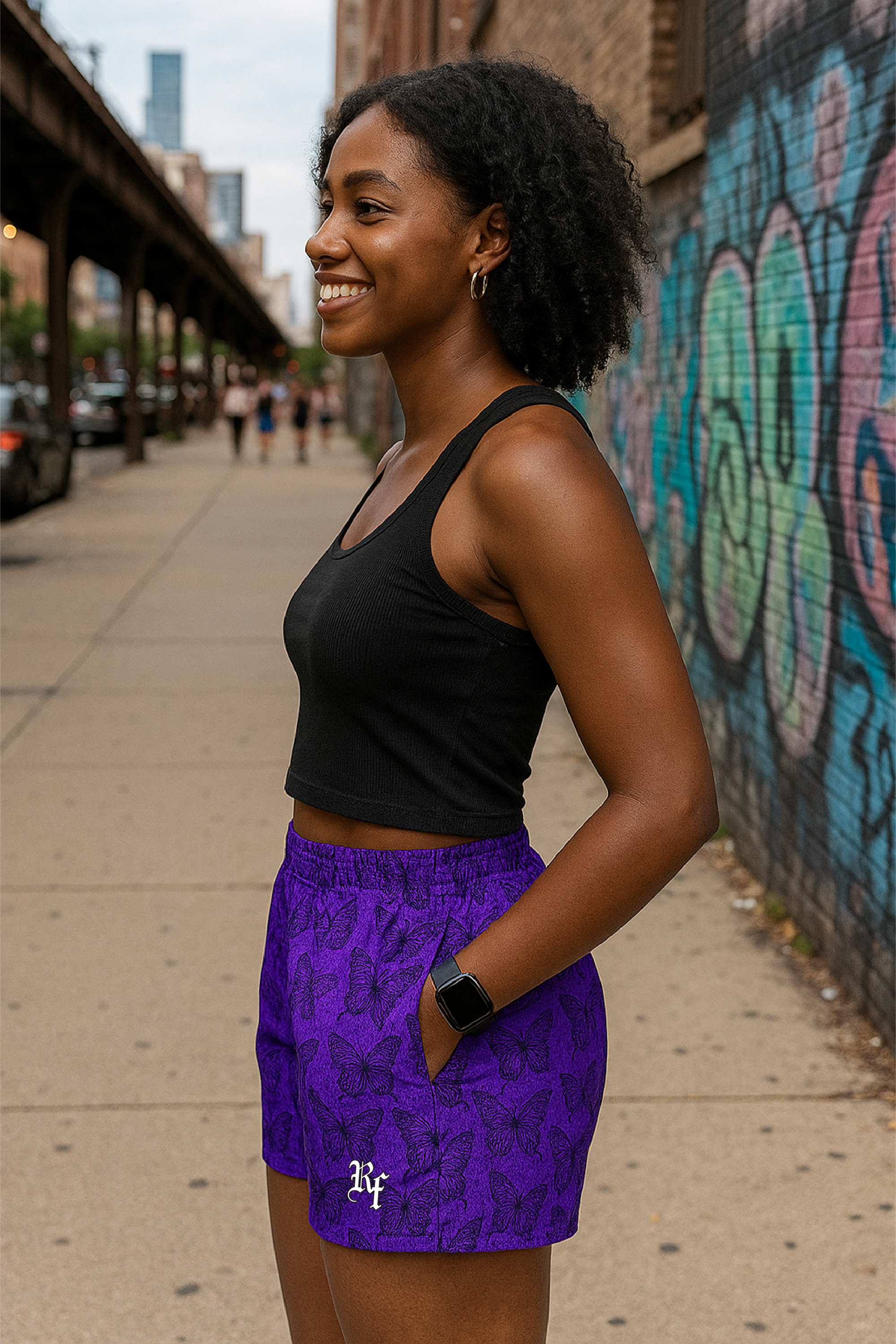 Woman in black top and purple shorts/black butterflies with a logo, standing on a sidewalk with graffiti in the background.