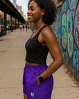 Woman in black top and purple shorts/black butterflies with a logo, standing on a sidewalk with graffiti in the background.