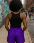 Women wearing a black tank top and purple shorts with black butterflies standing on a city street with graffiti.