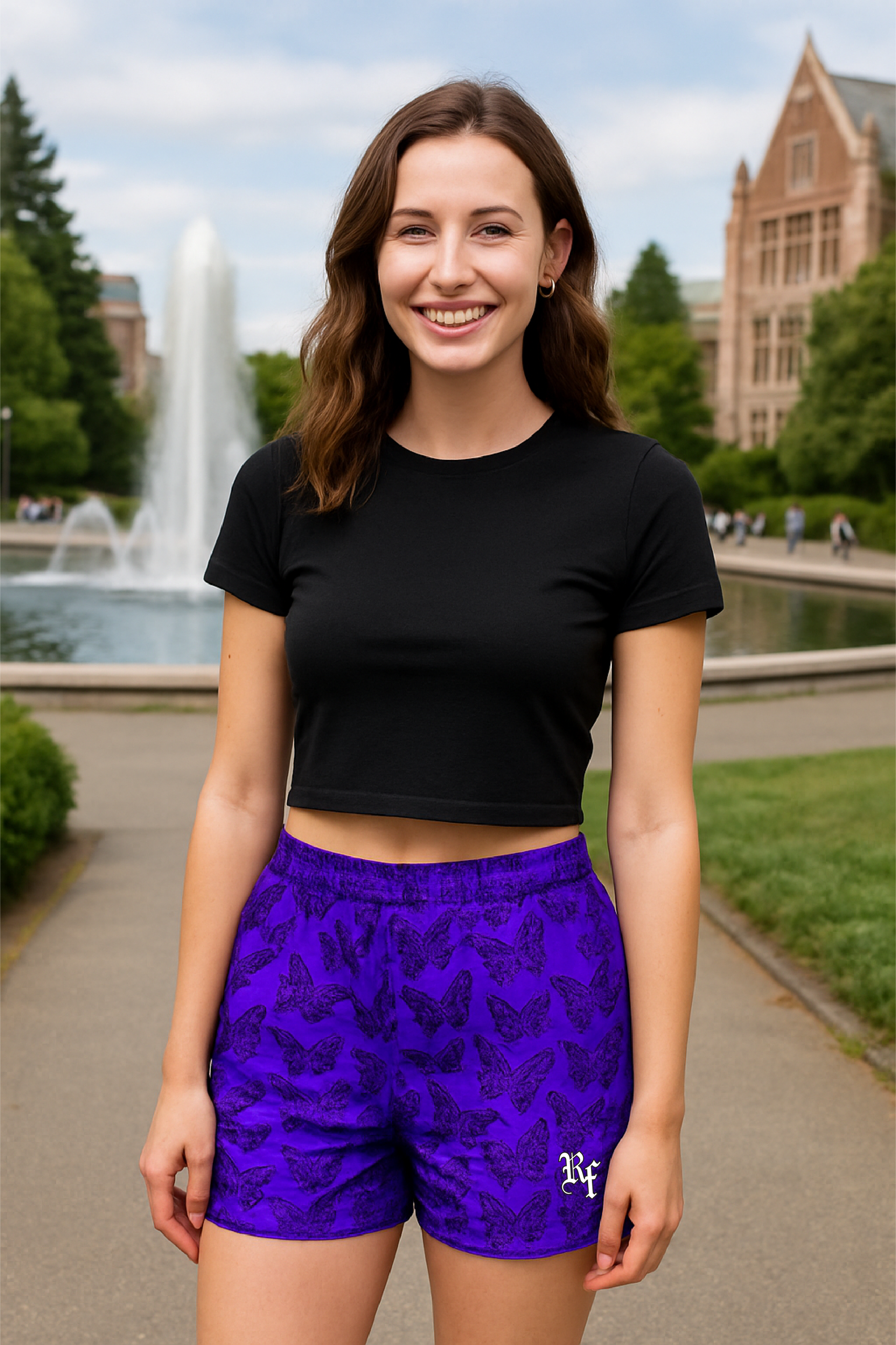 Woman wearing a black crop top and purple shorts with black butterflies, standing outdoors with a fountain and building in the background.