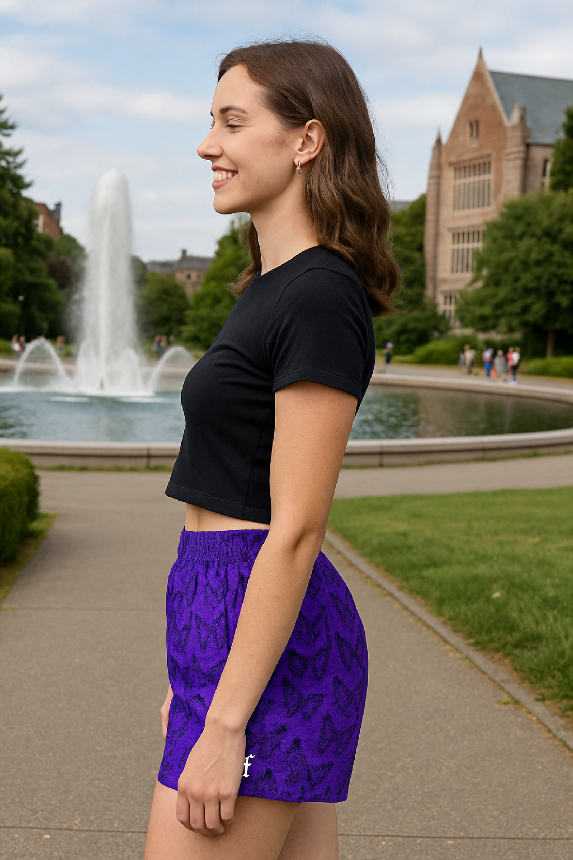 Woman wearing a black crop top and purple shorts with black butterflies, standing outdoors with a fountain and building in the background.