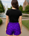 Woman wearing a black crop top and purple shorts with black butterflies, standing outdoors with a fountain and building in the background.