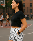 Woman standing on a basketball court wearing a black crop top and white shorts with black chain link pattern.