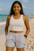 Woman in a white tank top and blue shorts with pink leaf pattern on a beach.