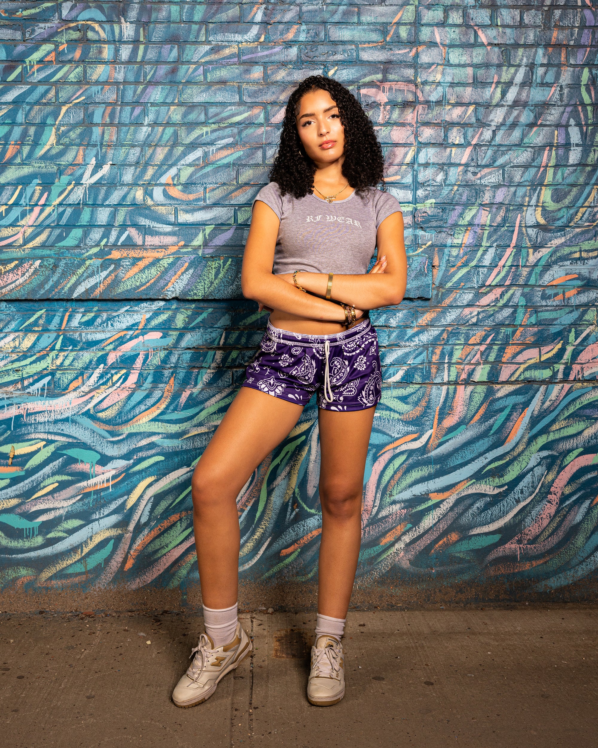 Woman standing near artful wall wearing RF-Wear Women's Paisley Shorts - Purple/White - Front View