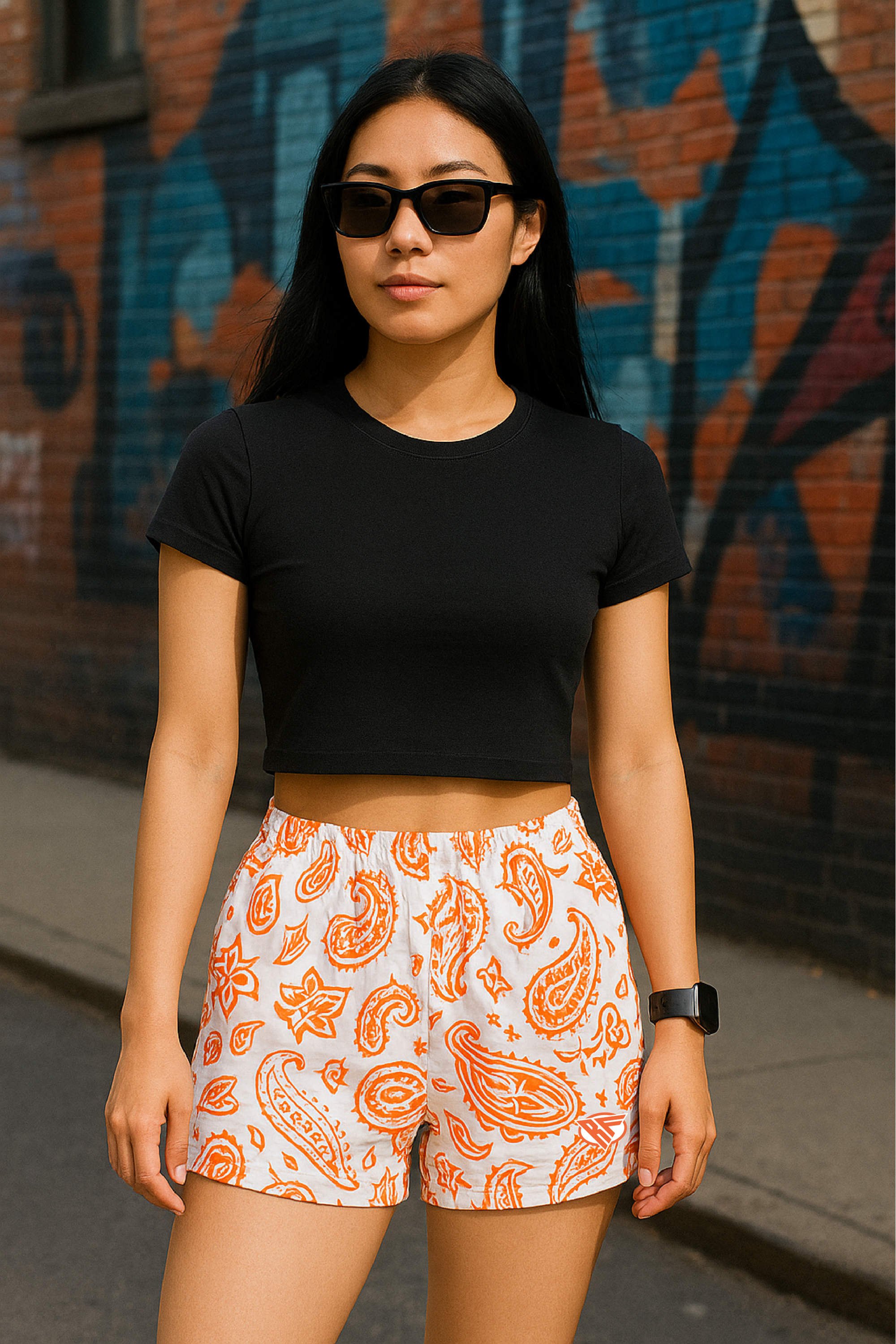 Woman wearing a black crop top and RF-Wear white/orange paisley shorts standing in front of a graffiti-covered wall.
