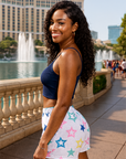 Woman in a blue top and colorful star shorts standing in front of a fountain and buildings.