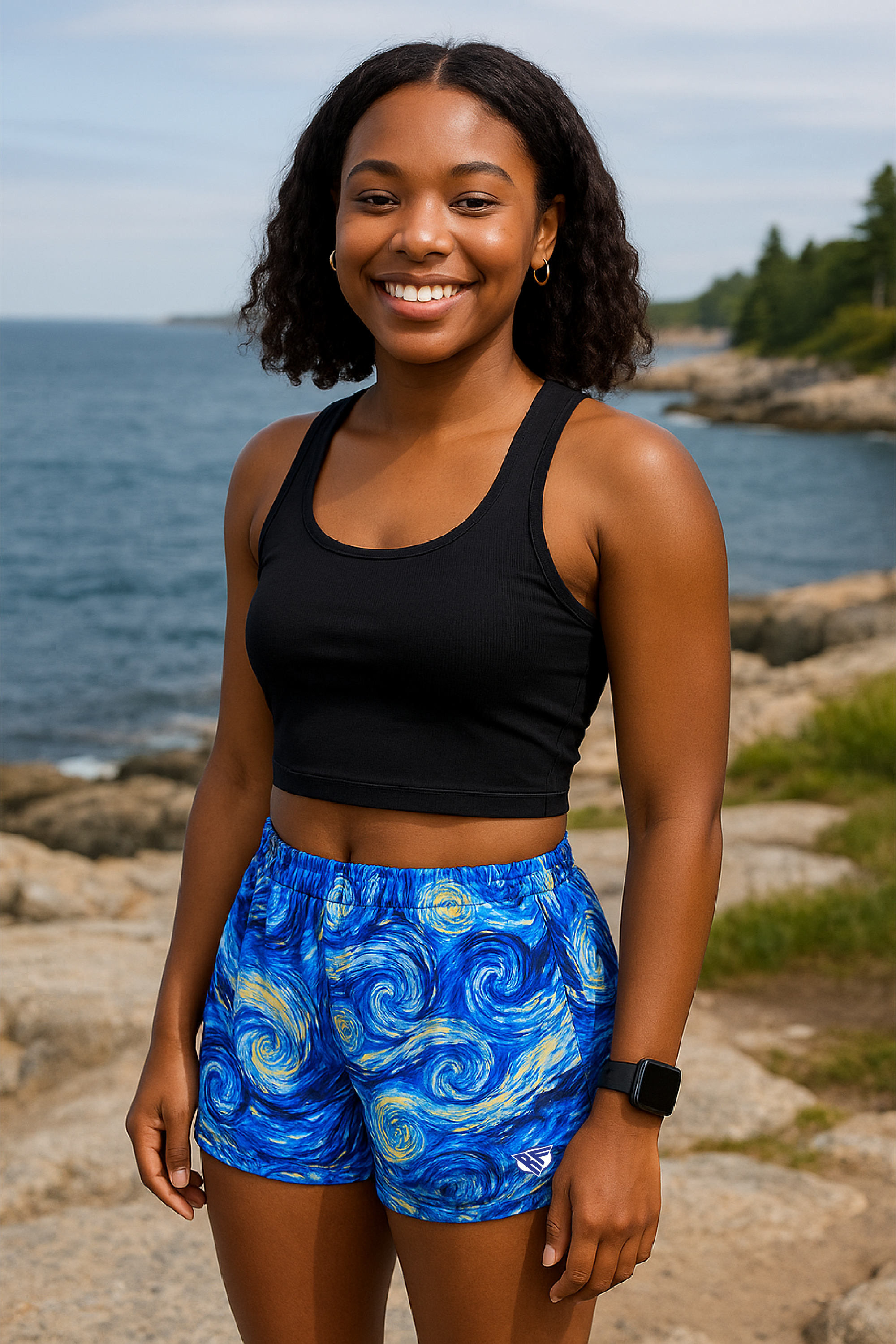 Woman in blue patterned shorts and black top standing on a path by the ocean with people in the background.