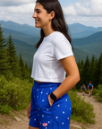 Woman in blue shorts with white star pattern and white shirt standing on a trail with mountains in the background