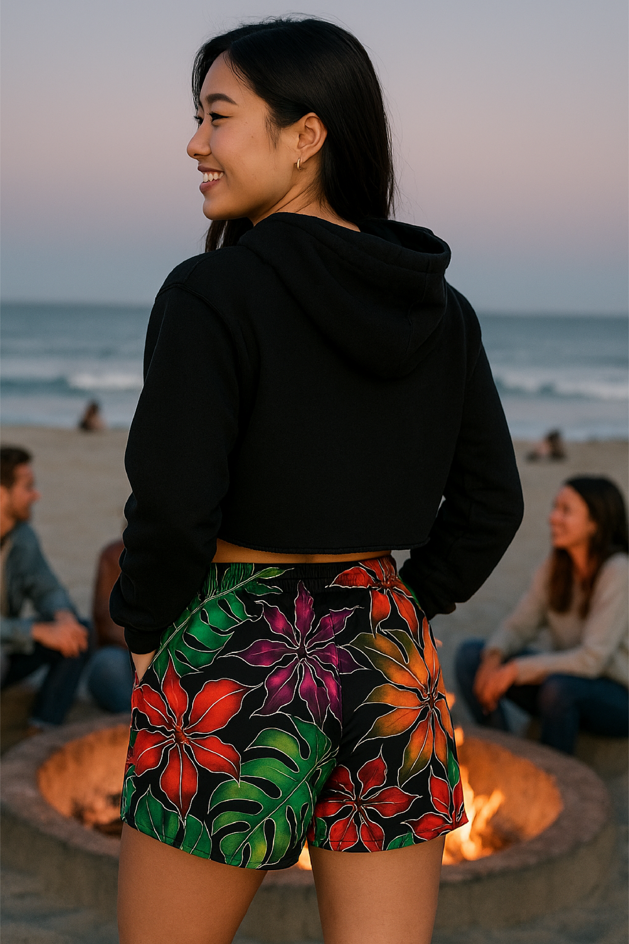 Person wearing a black hoodie and colorful shorts on a beach with people and a fire pit in the background.