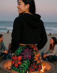 Person wearing a black hoodie and colorful shorts on a beach with people and a fire pit in the background.
