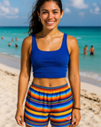 Woman in blue top and colorful shorts standing on a beach with ocean and people in the background