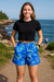 Woman wearing a black crop top and blue patterned shorts standing on a path near the ocean.