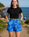 Woman wearing a black crop top and blue patterned shorts standing on a path near the ocean.