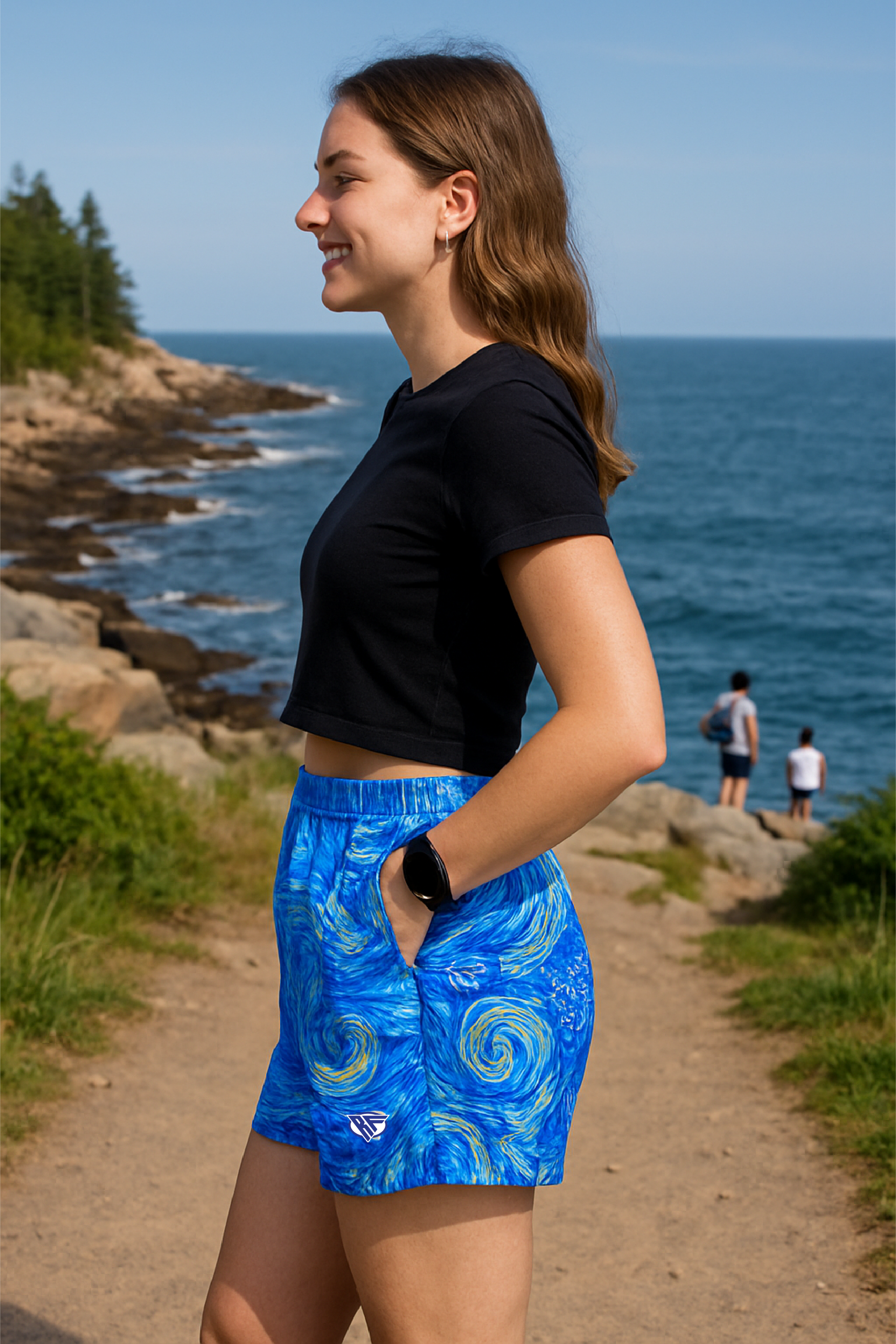 Woman in blue patterned shorts and black top standing on a path by the ocean with people in the background.