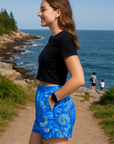 Woman in blue patterned shorts and black top standing on a path by the ocean with people in the background.